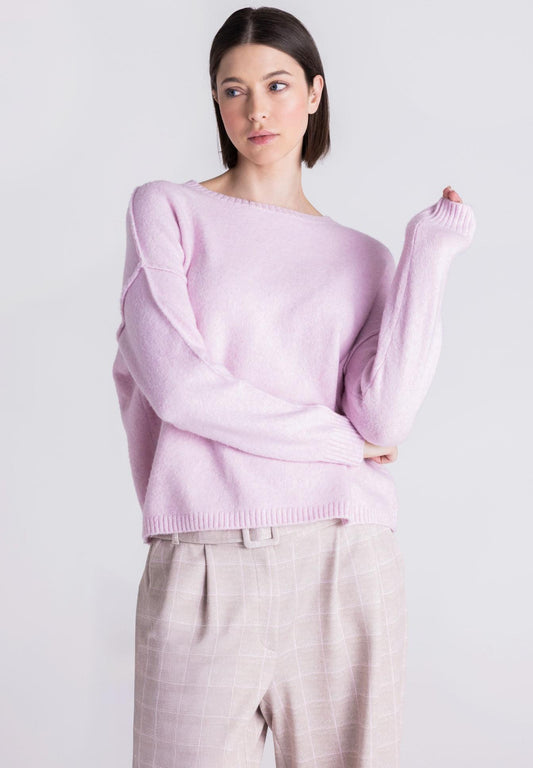 A woman modeling a light pink knit sweater with an asymmetrical draped neckline, paired with light beige plaid trousers, against a plain light gray background.