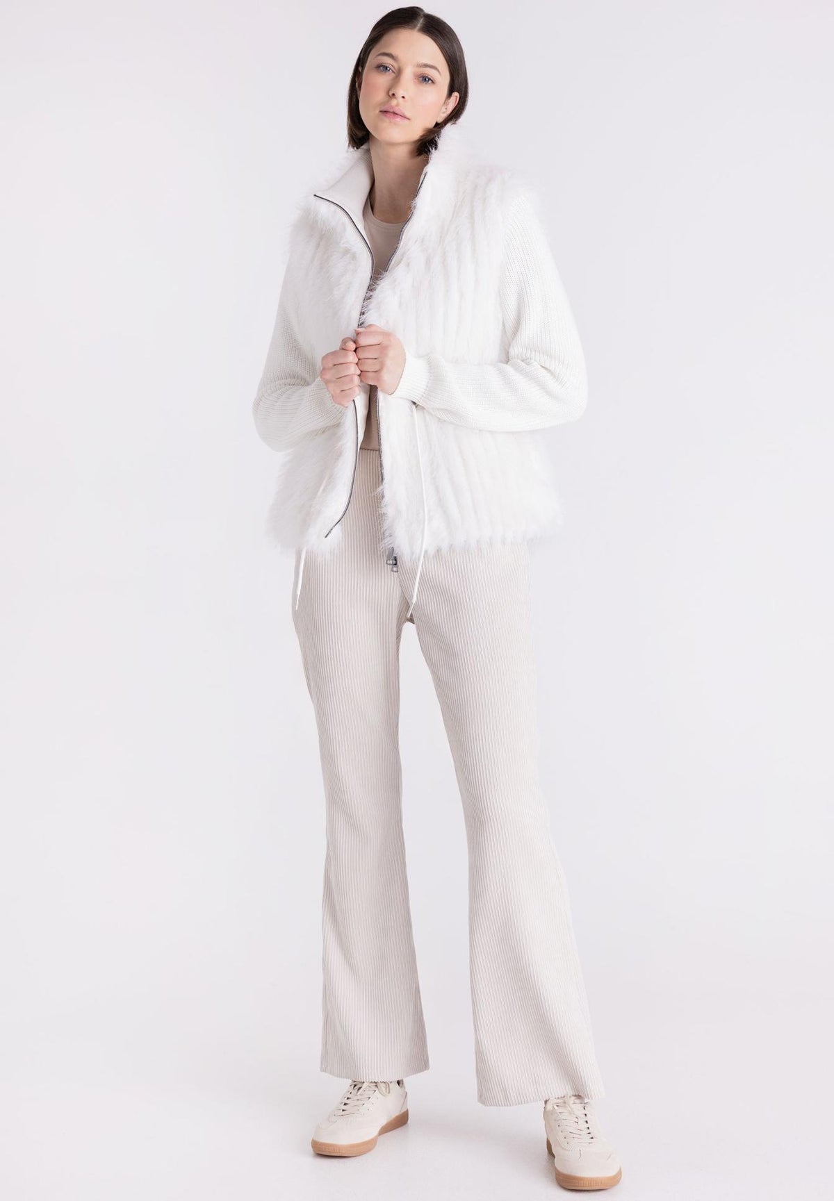 A woman modeling a white textured zip-up jacket with fringe trim, paired with light beige wide-leg trousers and white sneakers, against a plain white studio background.
