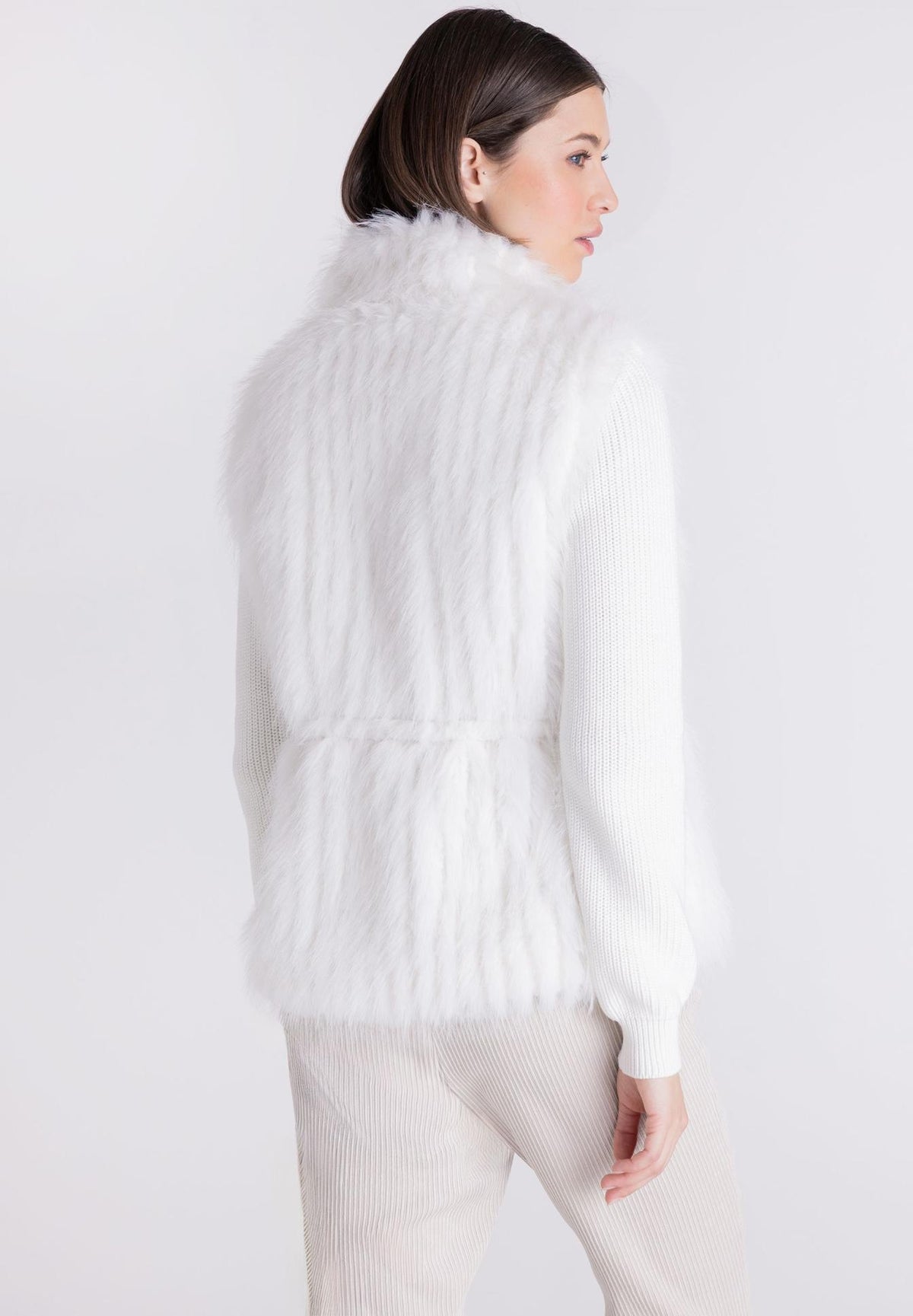 A woman shown from the back wearing a white faux fur vest over a white ribbed knit sweater and light beige trousers, photographed against a plain light gray studio background.