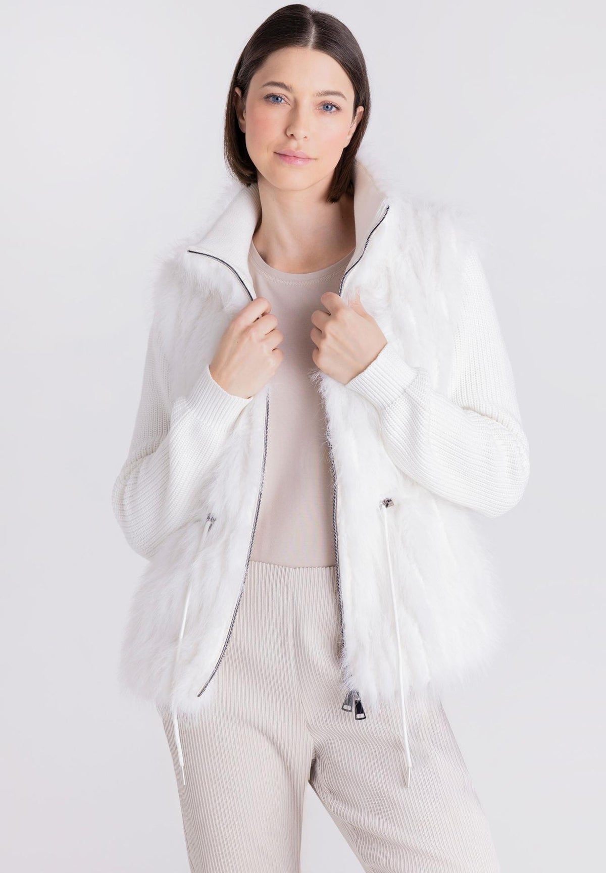 A woman modeling a white faux fur jacket with a high collar, worn over a beige ribbed top and matching beige pants, against a light gray studio background.