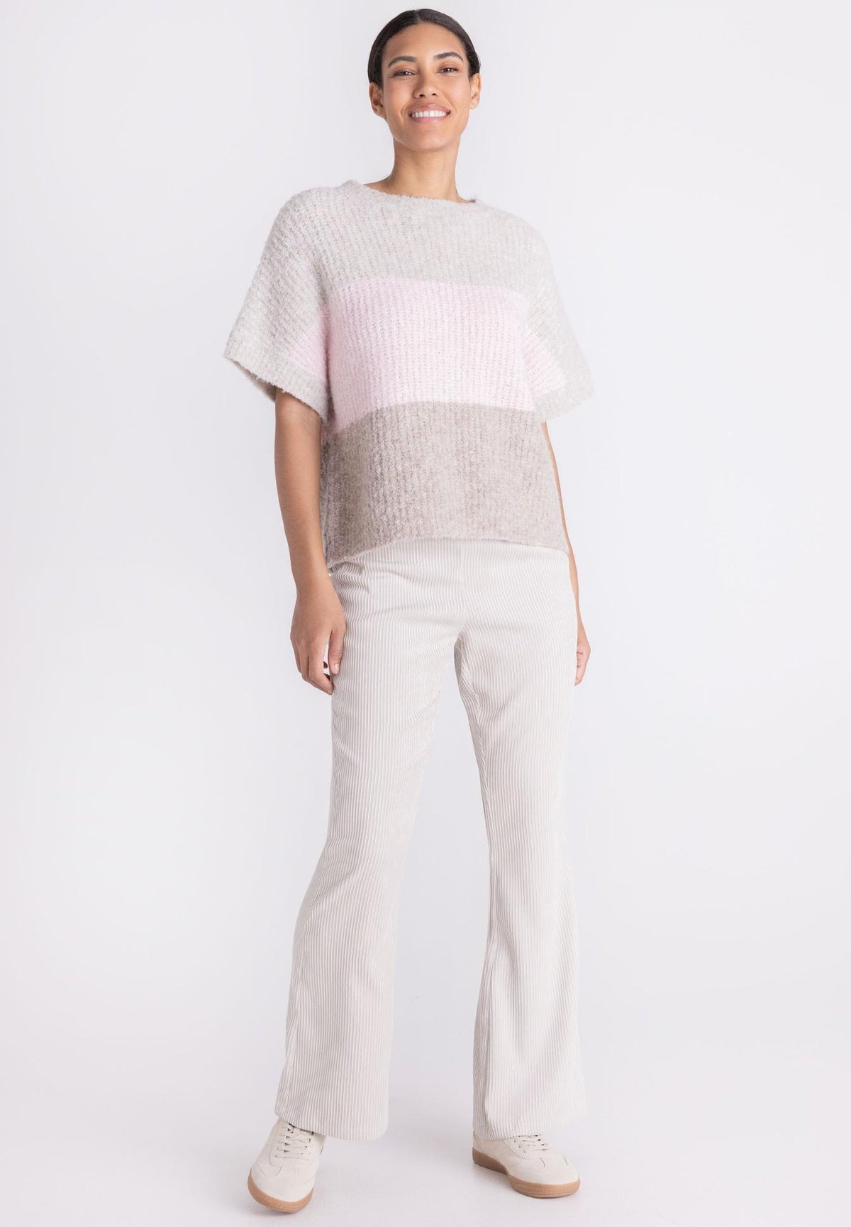A woman modeling a short-sleeve knit top with horizontal color-blocked stripes in light gray, pale pink, and taupe-beige, paired with light beige pants and white sneakers against a plain white studio background.