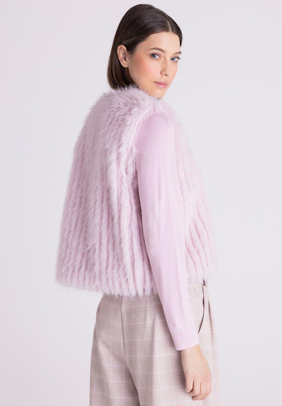 A woman wearing a light pink faux fur vest over a matching pink long-sleeve top and beige plaid trousers, photographed against a plain light gray studio background.
