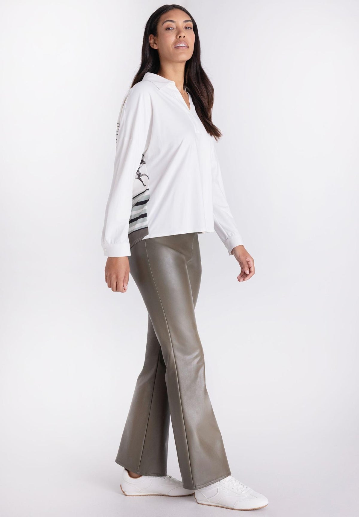 A woman modeling a white long-sleeve collared blouse with a black and white striped pattern on the lower sleeve, paired with metallic taupe pants, against a plain white studio background.