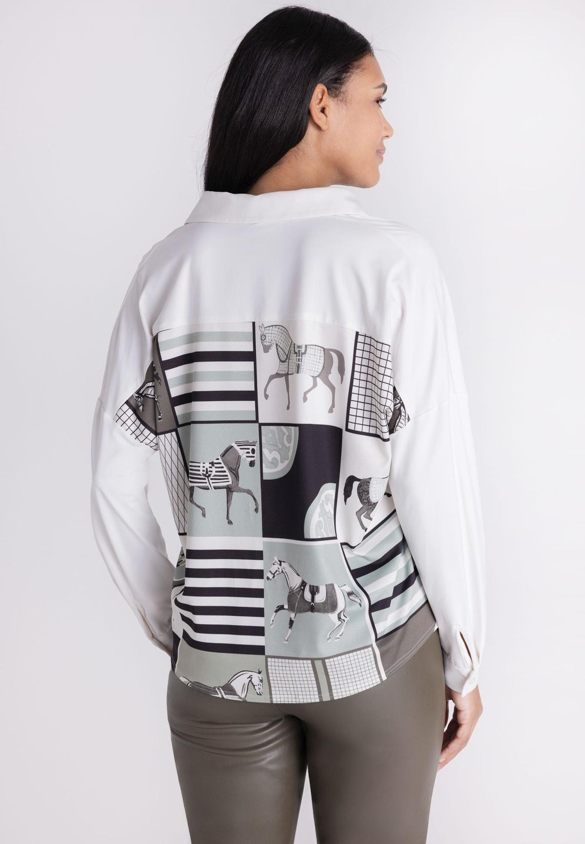A woman shown from the back wearing a white long-sleeve shirt with a large patchwork horse print on the back in black, gray, and white, paired with gray pants, against a light gray studio background.