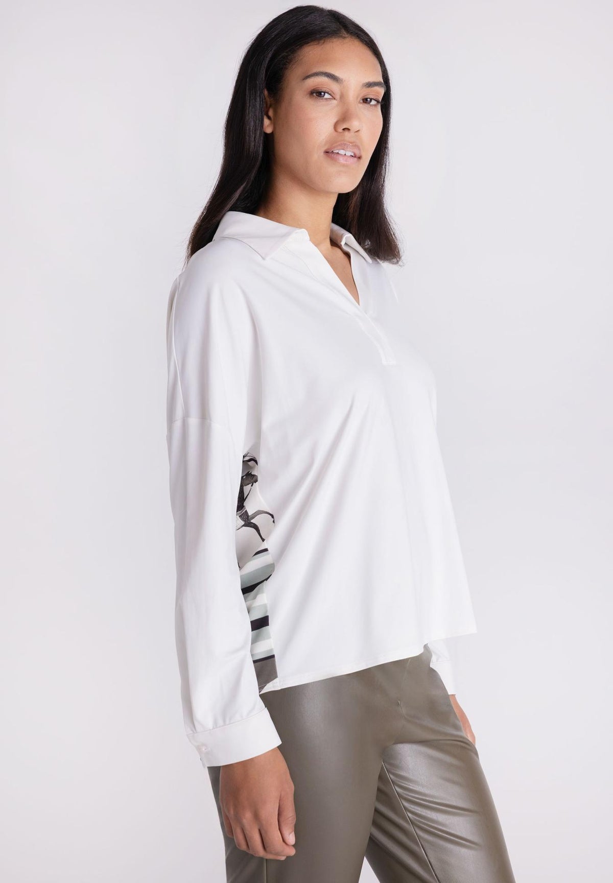 A woman modeling a white long-sleeve collared blouse with a black and white striped pattern on the lower sleeve, paired with metallic taupe pants, against a plain white studio background.