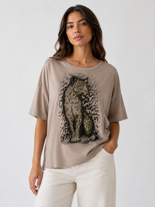 A woman modeling a taupe short-sleeve t-shirt with a large graphic print of a leopard on the front, paired with off-white pants, against a plain light gray background.