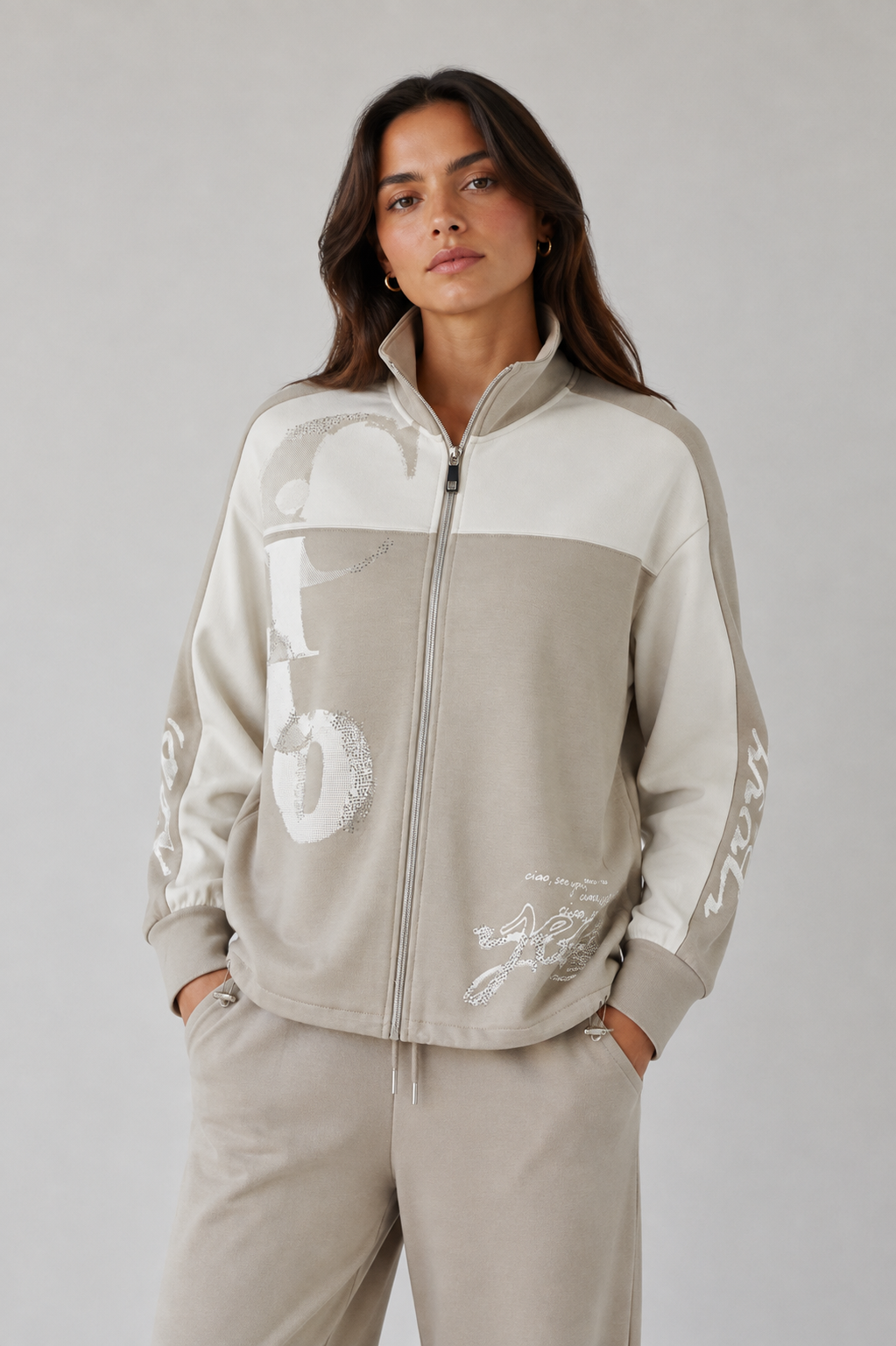A woman modeling a beige and off-white color-blocked zip-up track jacket with graphic lettering on the front and sleeves, paired with matching beige sweatpants, against a neutral gray studio background.