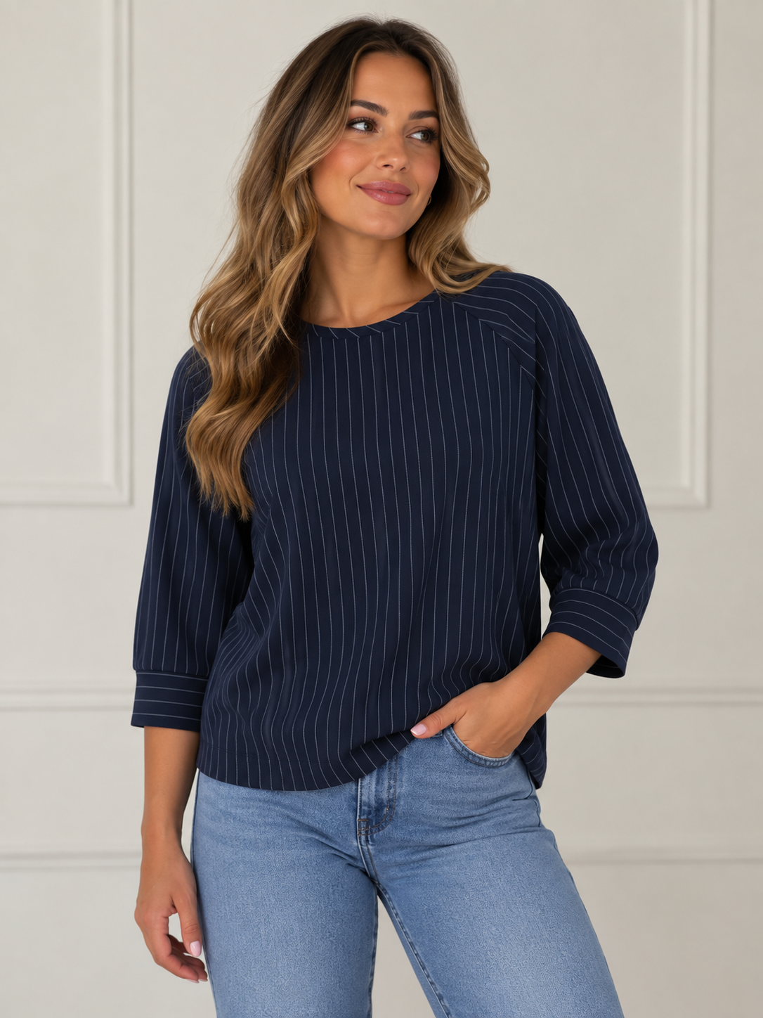 A woman wearing a navy blue long-sleeve top with thin white vertical pinstripes and light blue jeans, standing against a white paneled wall.