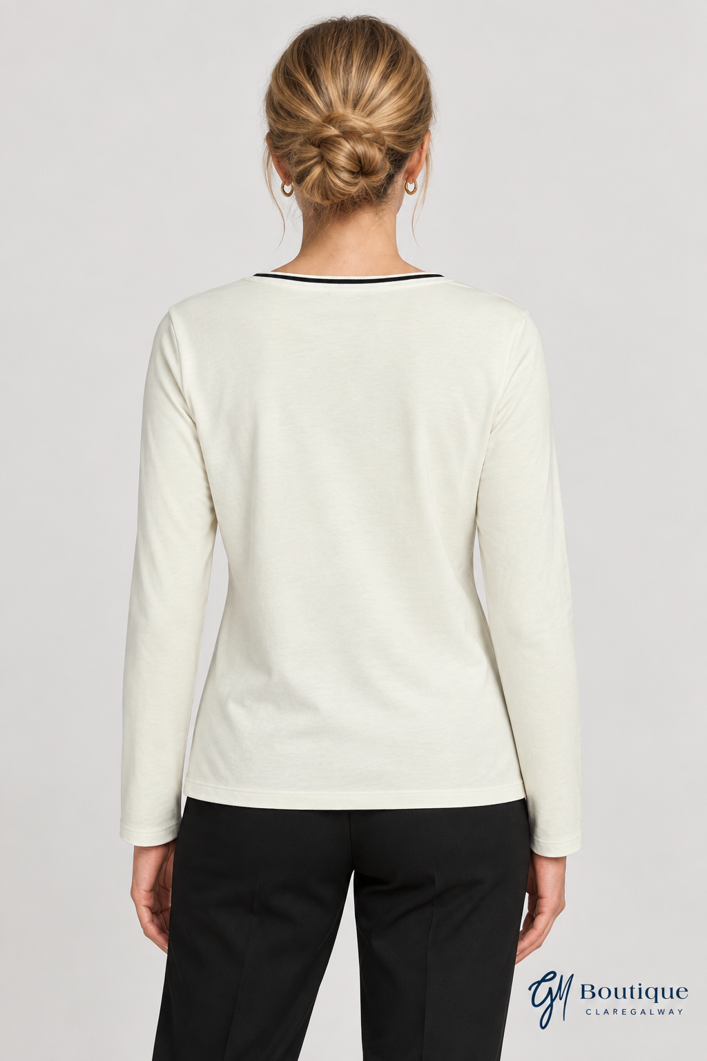 A woman viewed from the back wearing a cream long-sleeve knit top with thin black trim at the neckline, paired with black pants, against a light gray studio background.