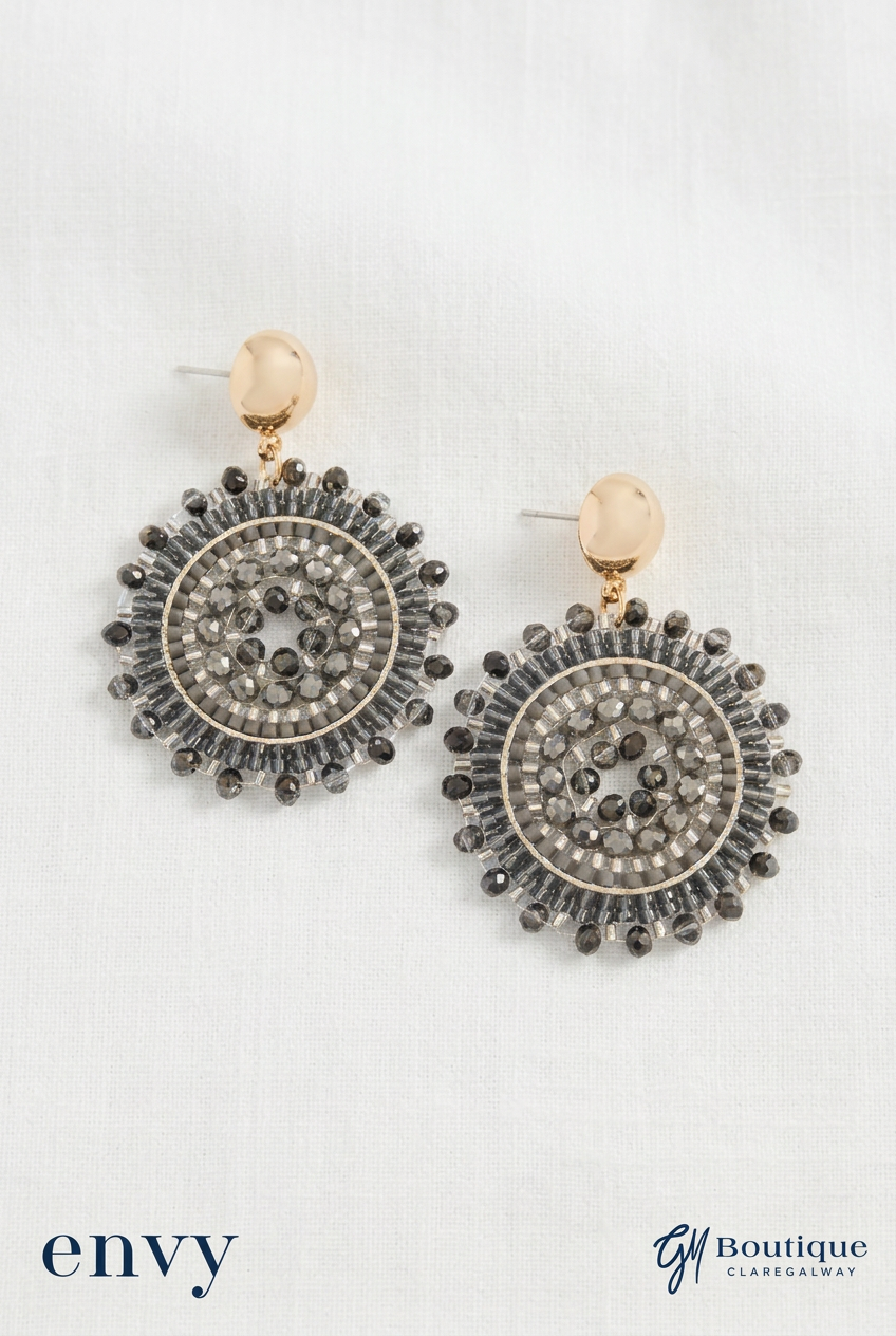 A pair of round drop earrings with gold-toned posts and circular dangles featuring dark gray and clear rhinestone patterns, displayed on a white textured fabric background with 'envy' and 'Gm Boutique claregalway' text at the bottom.