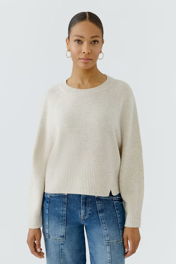 A woman modeling a light cream knit crewneck sweater with raglan sleeves, paired with blue jeans, against a light gray studio background.