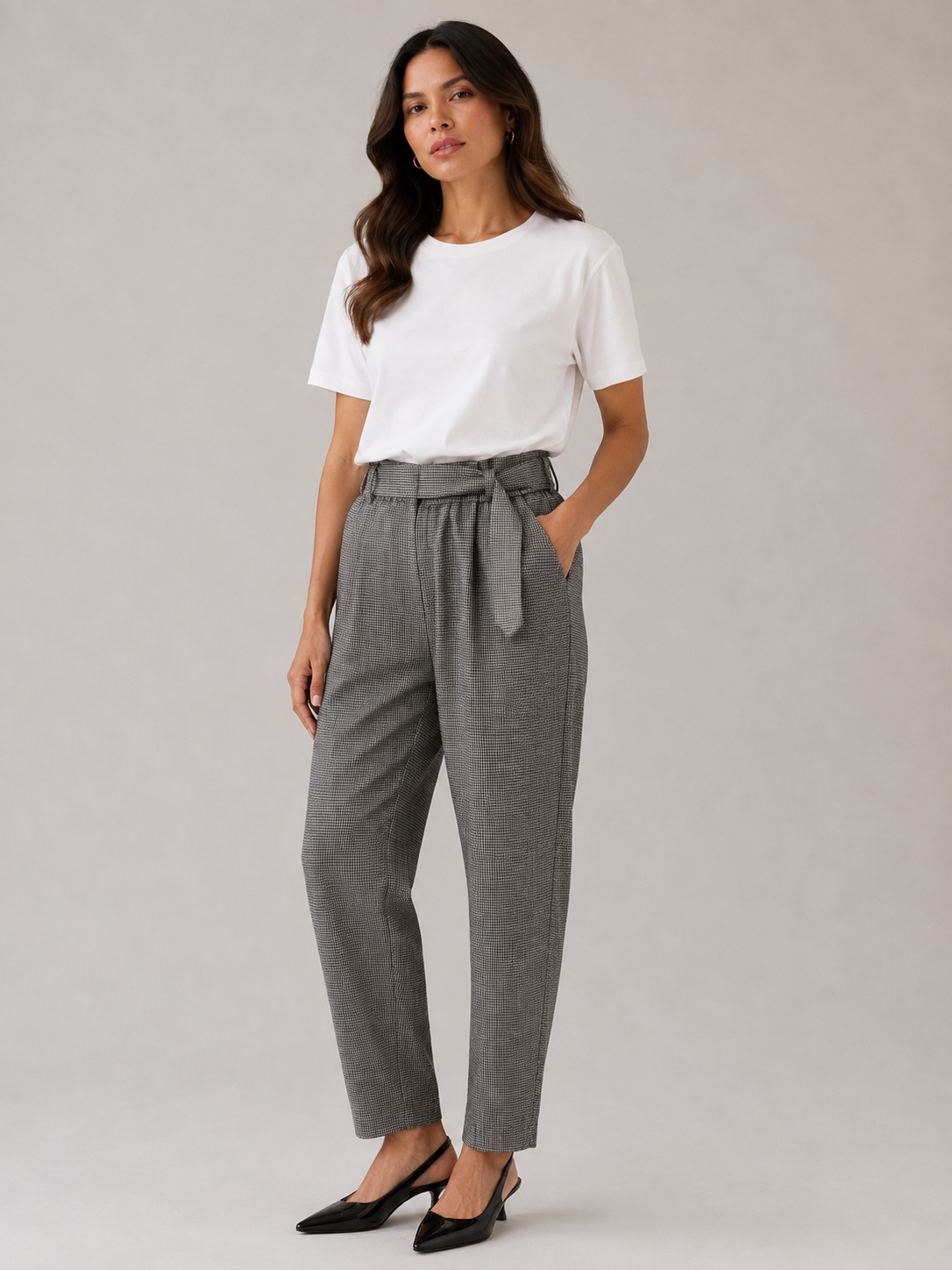 A woman modeling high-waisted gray trousers with a tie waist, paired with a white t-shirt and black heels, against a neutral studio background.