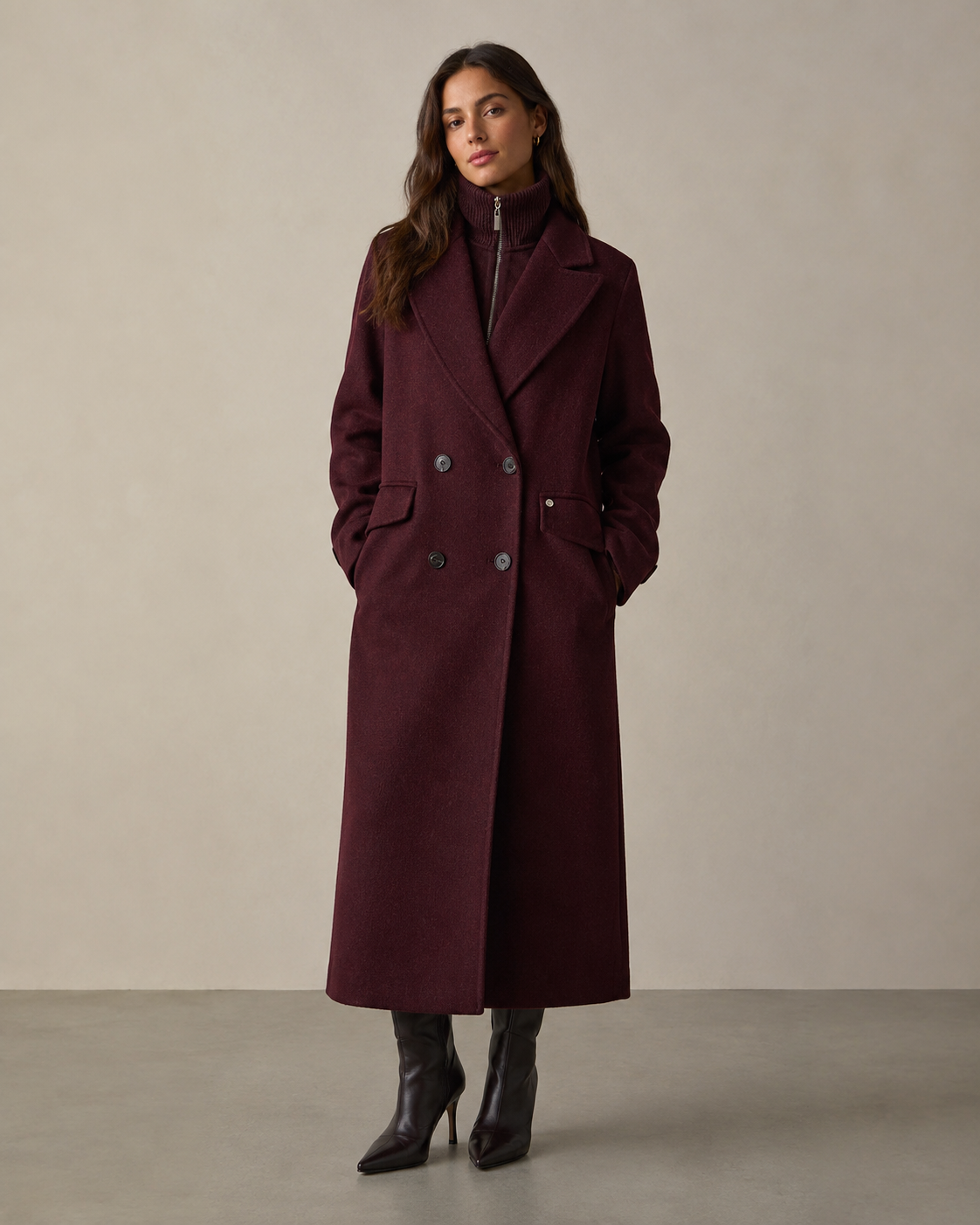 A woman modeling a long, double-breasted wool coat in deep burgundy, paired with black leather ankle boots, against a neutral beige studio background.