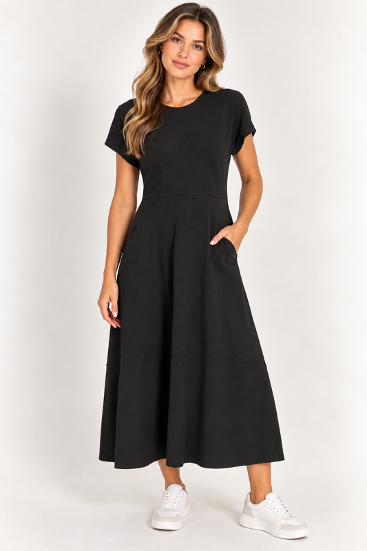 A woman modeling a black short-sleeve midi dress with a flared skirt and side pockets, paired with white sneakers, against a plain light gray studio background.