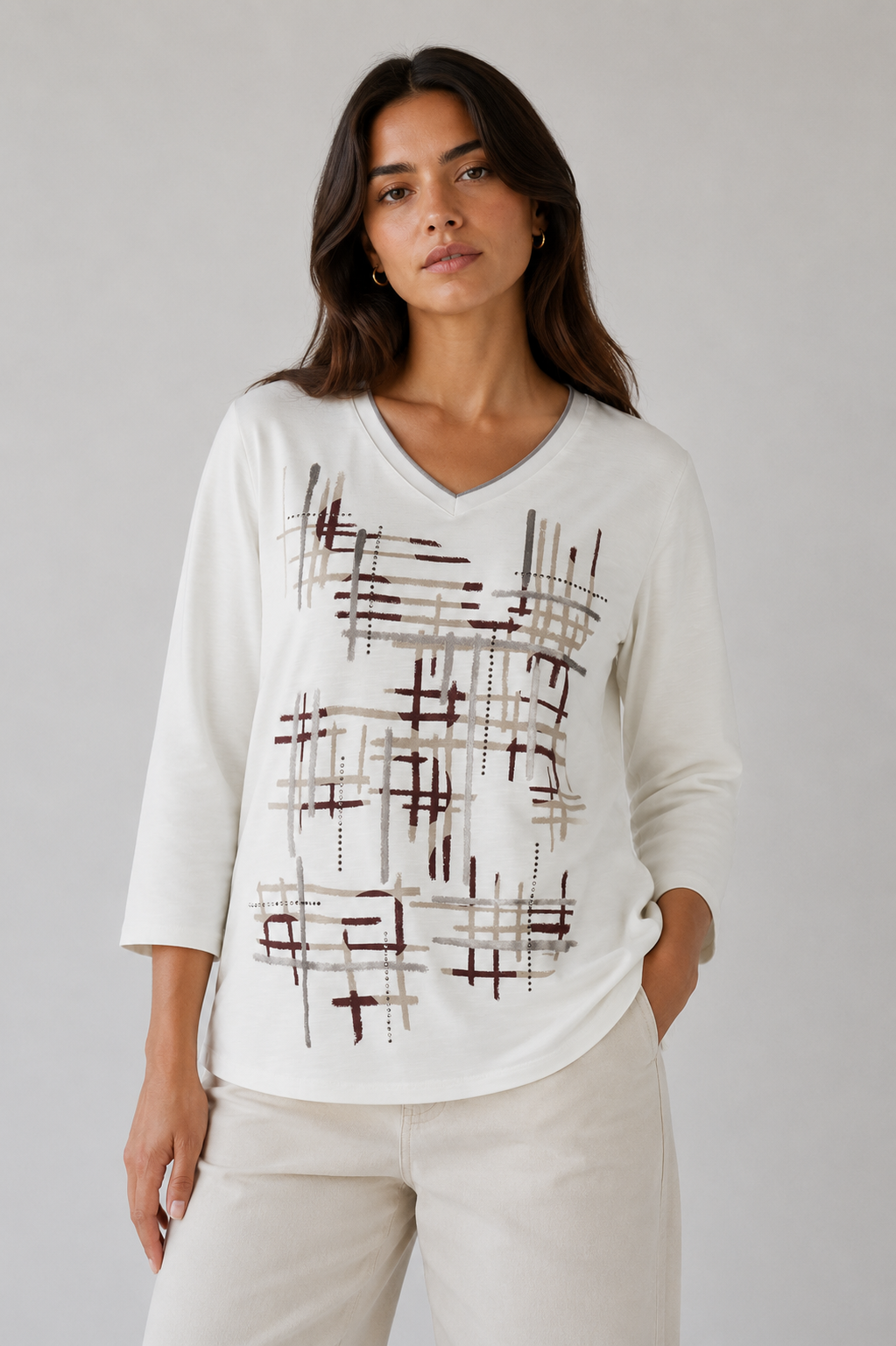 A woman modeling a cream-colored long-sleeve V-neck top with an abstract plaid pattern in brown and gray, paired with cream pants, against a neutral gray background.