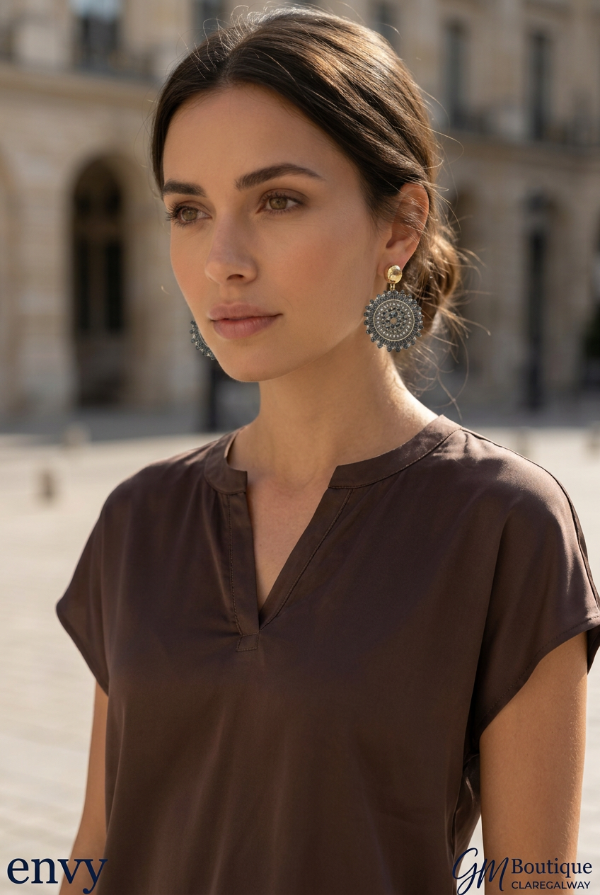 A woman wearing a brown short-sleeve V-neck top and large circular silver earrings, photographed outdoors in front of a blurred stone building. The image includes 'envy' and 'GM Boutique' watermarks.