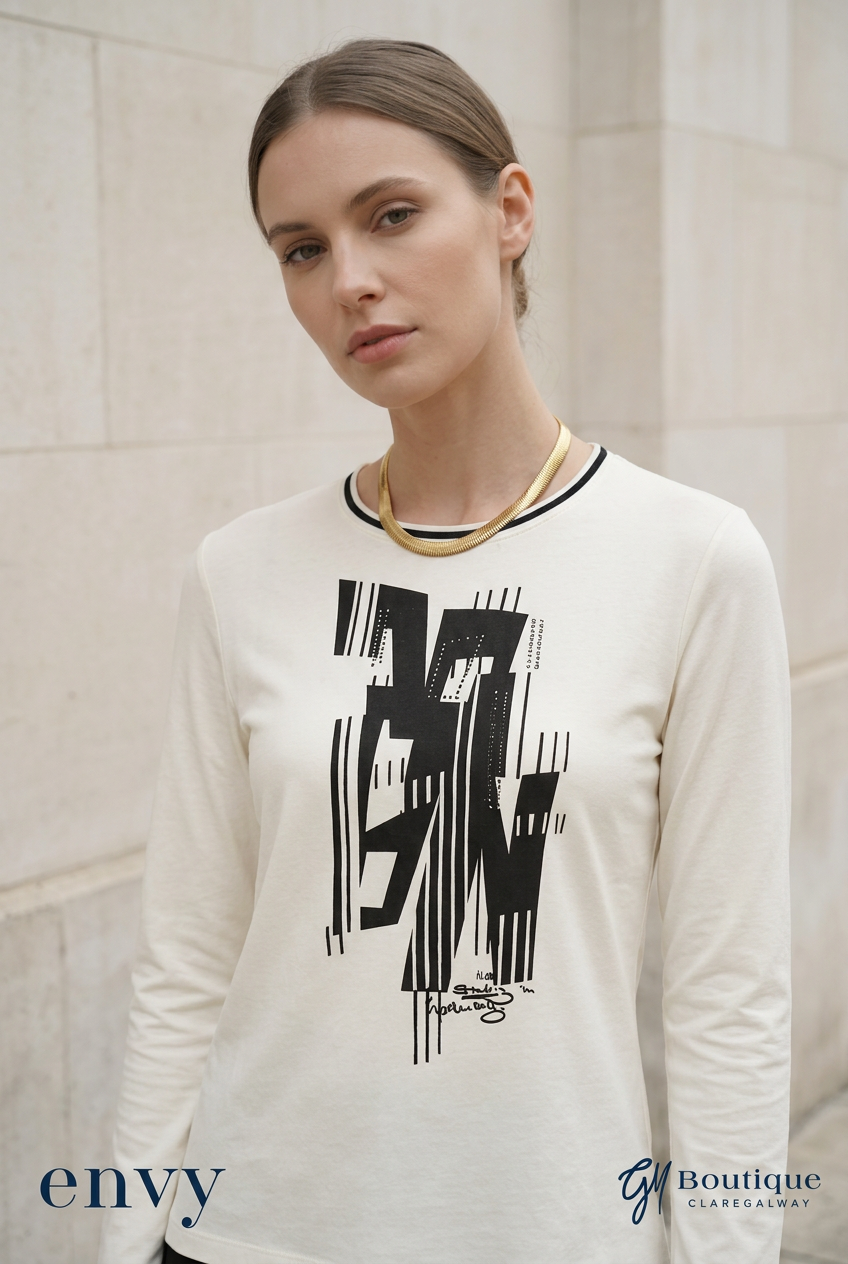 A woman wearing Gm boutique  cream-colored long-sleeve t-shirt with a black graphic print on the front, styled with a gold chain necklace, set against a light stone wall background.