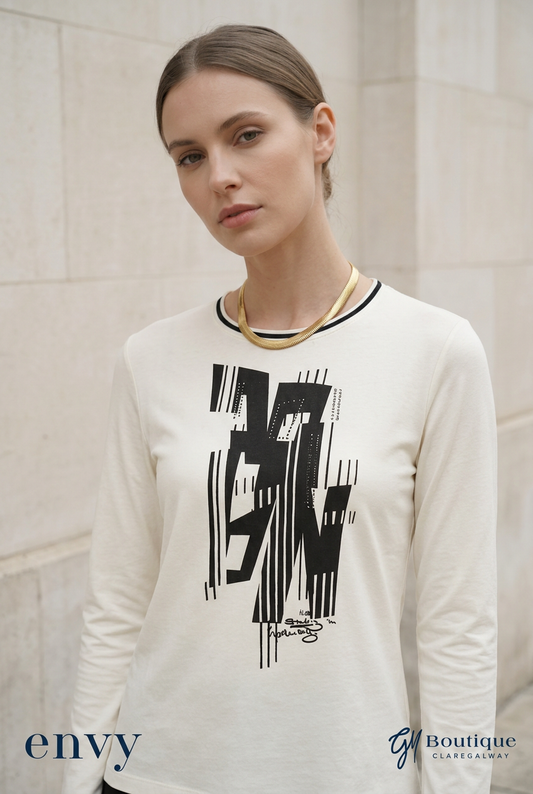 A woman wearing Gm boutique  cream-colored long-sleeve t-shirt with a black graphic print on the front, styled with a gold chain necklace, set against a light stone wall background.