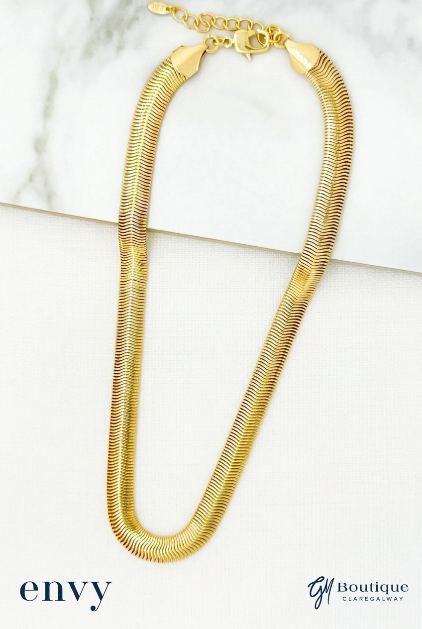 A gold snake chain necklace draped in a U-shape on a white marble slab and white surface, with 'envy' and 'GM BOUTIQUE ' text at the bottom.
