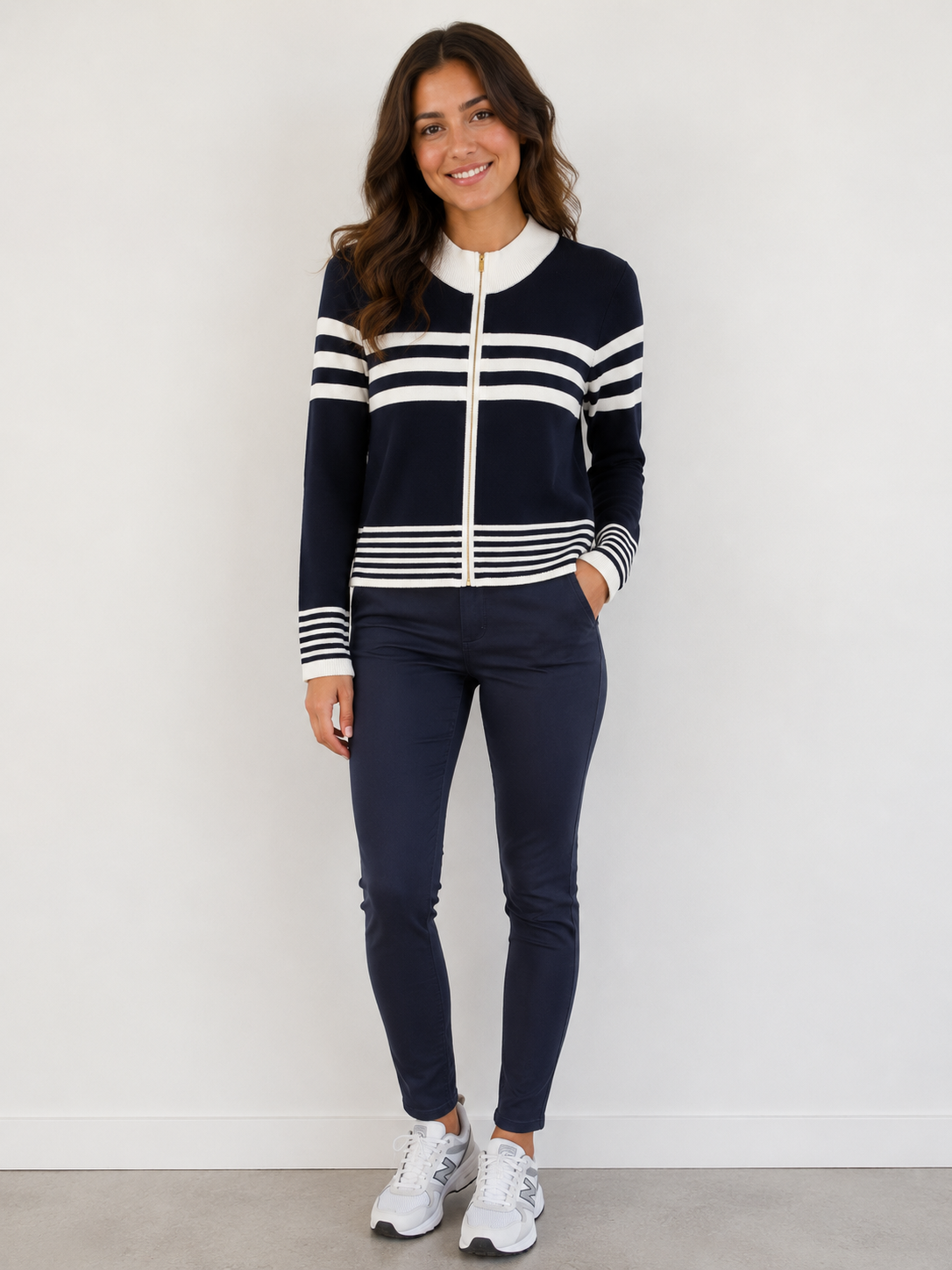 A woman modeling a navy blue zip-up cardigan with white stripes and a white collar, paired with navy pants and white sneakers, standing against a plain white wall.
