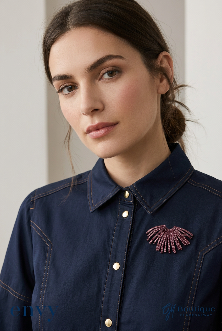 A close-up of a woman wearing a dark navy denim shirt with gold buttons and a pink beaded fan-shaped brooch on the chest. The photo has a soft, warm-toned aesthetic with a blurred background.