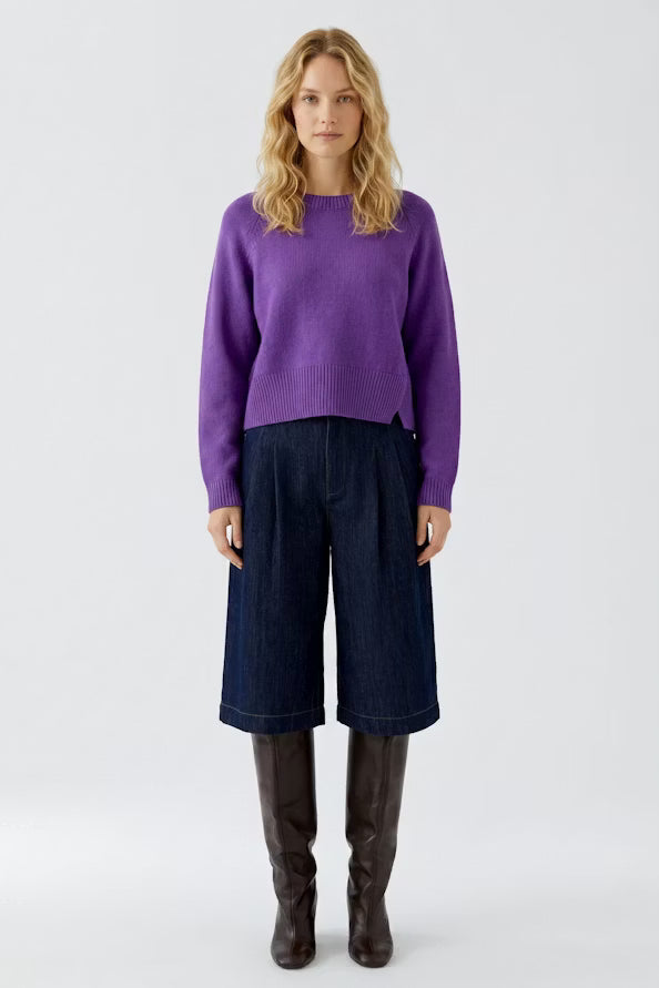 A woman stands against a light gray studio background wearing a purple knit sweater, dark blue wide-leg cropped pants, and dark brown knee-high boots.