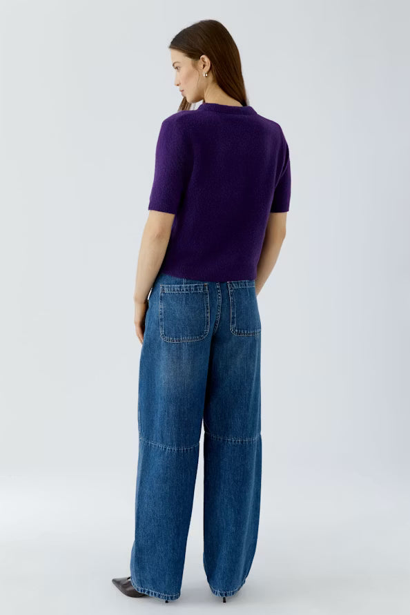 A woman is shown from the back wearing a short-sleeve purple knit top and wide-leg blue denim jeans against a light gray studio background.