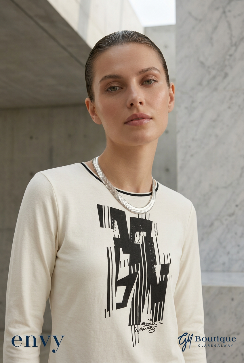A woman wearing Gm Boutique cream-colored long-sleeve crewneck sweatshirt with a bold black graphic print on the front, set against a modern concrete architectural background.