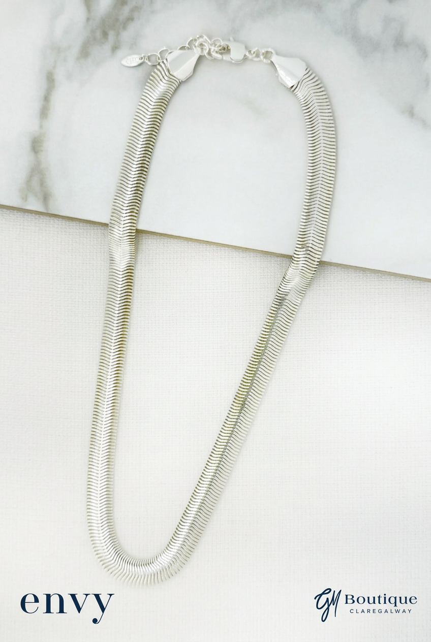 A silver snake chain necklace with a lobster clasp, displayed on a white textured surface with a marble slab in the background. The brand logos 'envy' and 'GM BOUTIQUE CLAREGALWAY' are visible in the bottom corners.