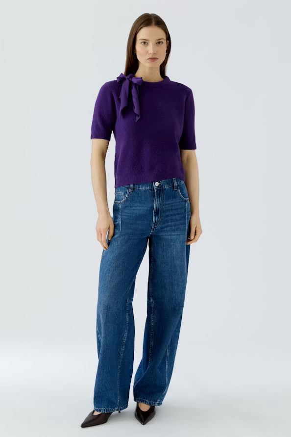 A woman modeling a short-sleeve purple knit top with a bow detail at the neckline, paired with wide-leg blue jeans and dark pointed shoes, against a light gray studio background.
